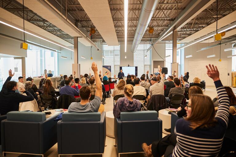 Lecture set up with a group of 50 people, some with their hands raised