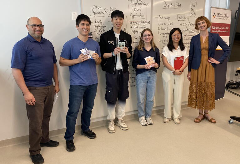 Six people standing in front of a whiteboard, 3 are holding awards
