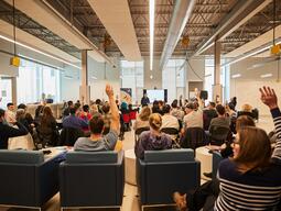 Lecture set up with a group of 50 people, some with their hands raised