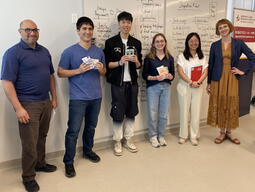 Six people standing in front of a whiteboard, 3 are holding awards